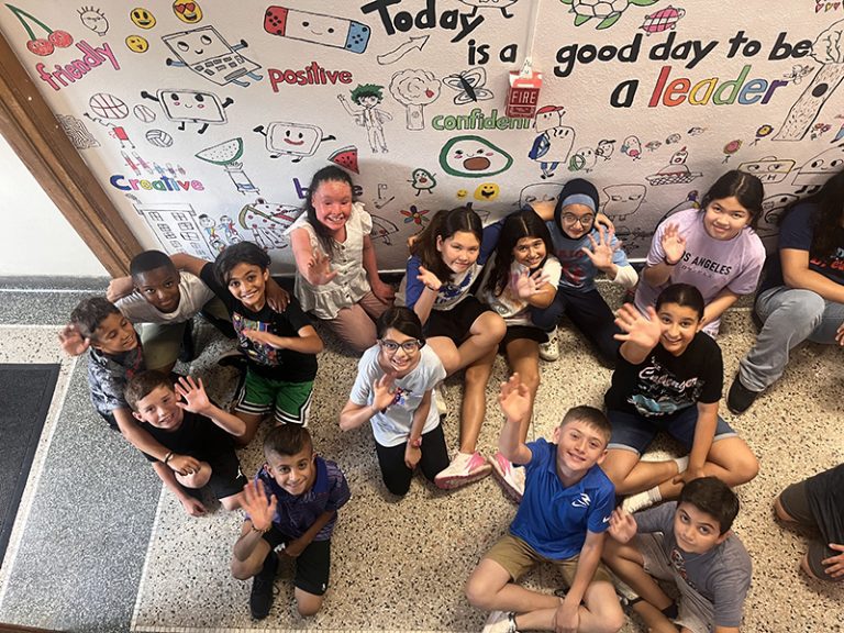 A group of students in the Lindbergh Elementary 2024 Summer Discovery Program pose in front of a mural they are creating at the school.